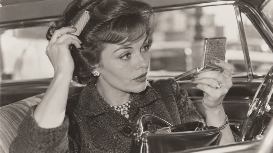 A woman sits in a car, holding a compact mirror in one hand and combing her hair with the other. She appears well-dressed, wearing earrings and a necklace, with an open handbag on her lap. The photo is in black and white.