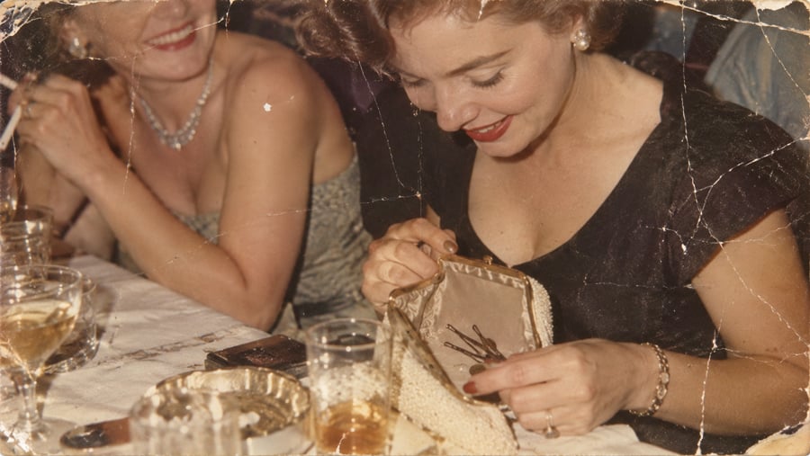Two women sit at a table with drinks and an ashtray. One woman, smiling, opens a clutch purse. The photo has a vintage look with visible creases and wear.