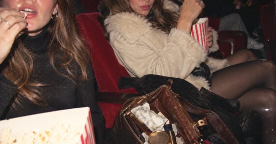 Two people sit in red theater seats eating popcorn. One holds a handbag with various items visible. Both are dressed in dark clothes; one wears a fur coat. The background is dim, suggesting they are inside a movie theater.