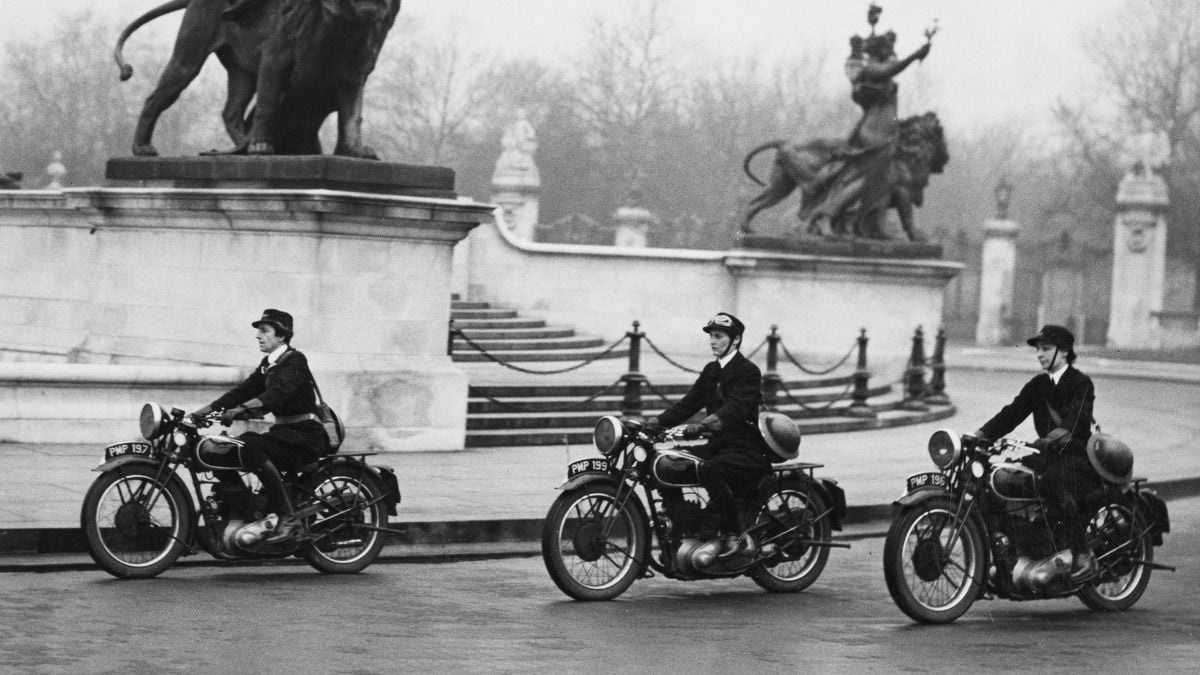 Three uniformed women ride vintage motorcycles near a large monument with statues of lions and figures, on a foggy day. The scene appears to be historical and set in an urban park or plaza.