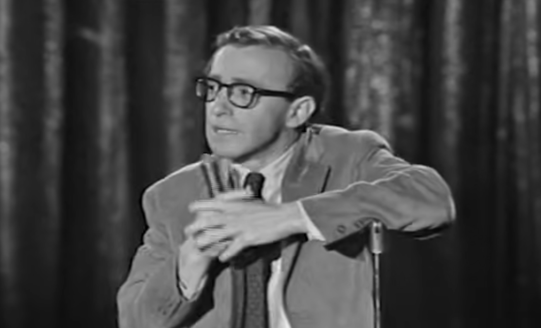 Woody Allen wearing glasses and a suit jacket, holding a microphone while performing on stage.