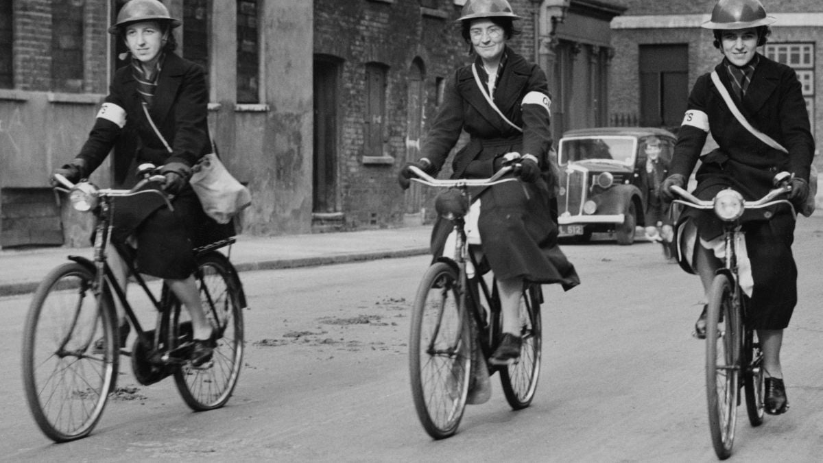 Three women wearing uniforms and helmets ride bicycles on a city street, each with a satchel and an armband. Old buildings and a vintage car are visible in the background. The photo appears to be from the mid-20th century.