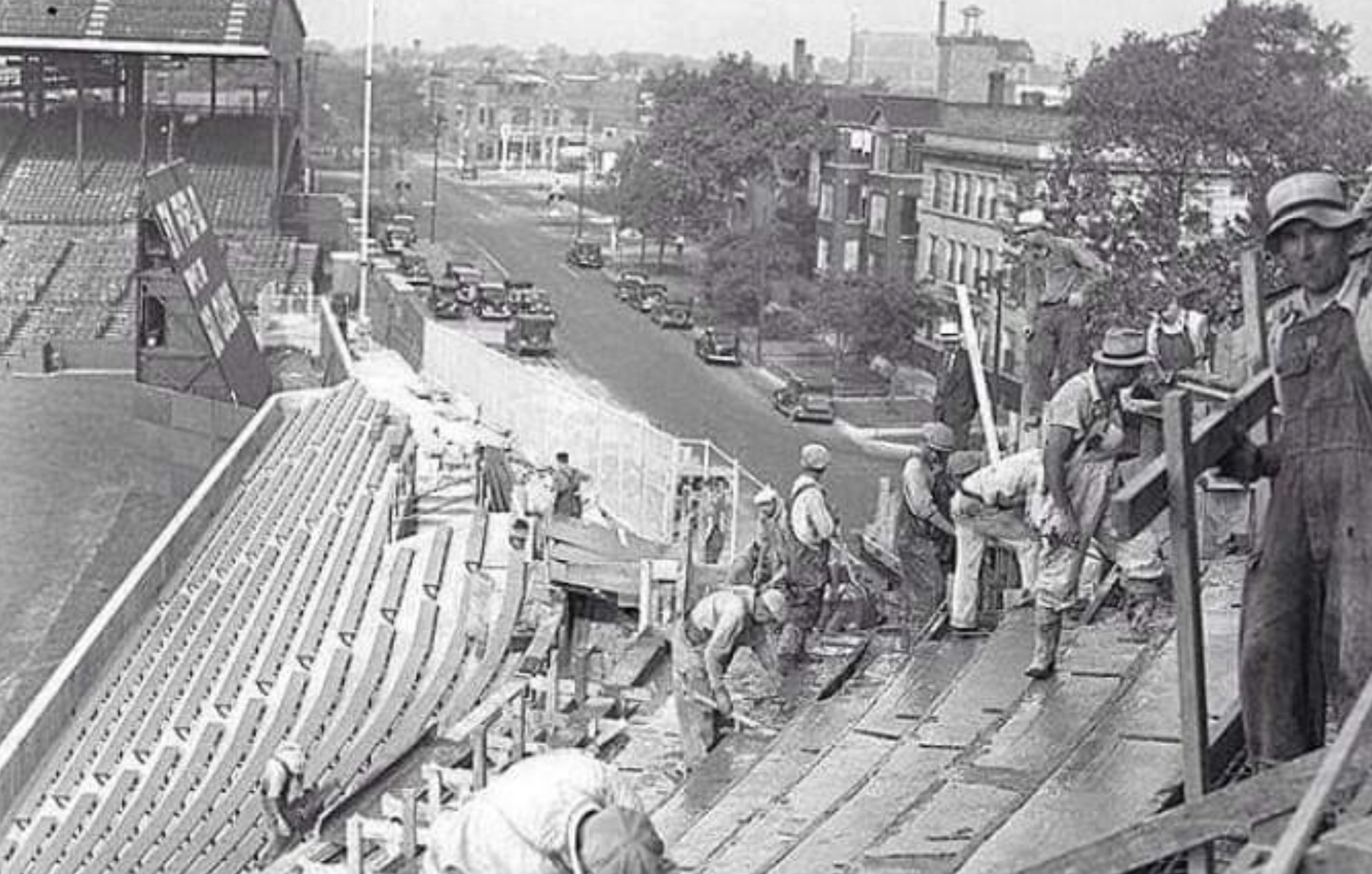 Construction workers installing wooden stadium seating in a ballpark overlooking a city street.