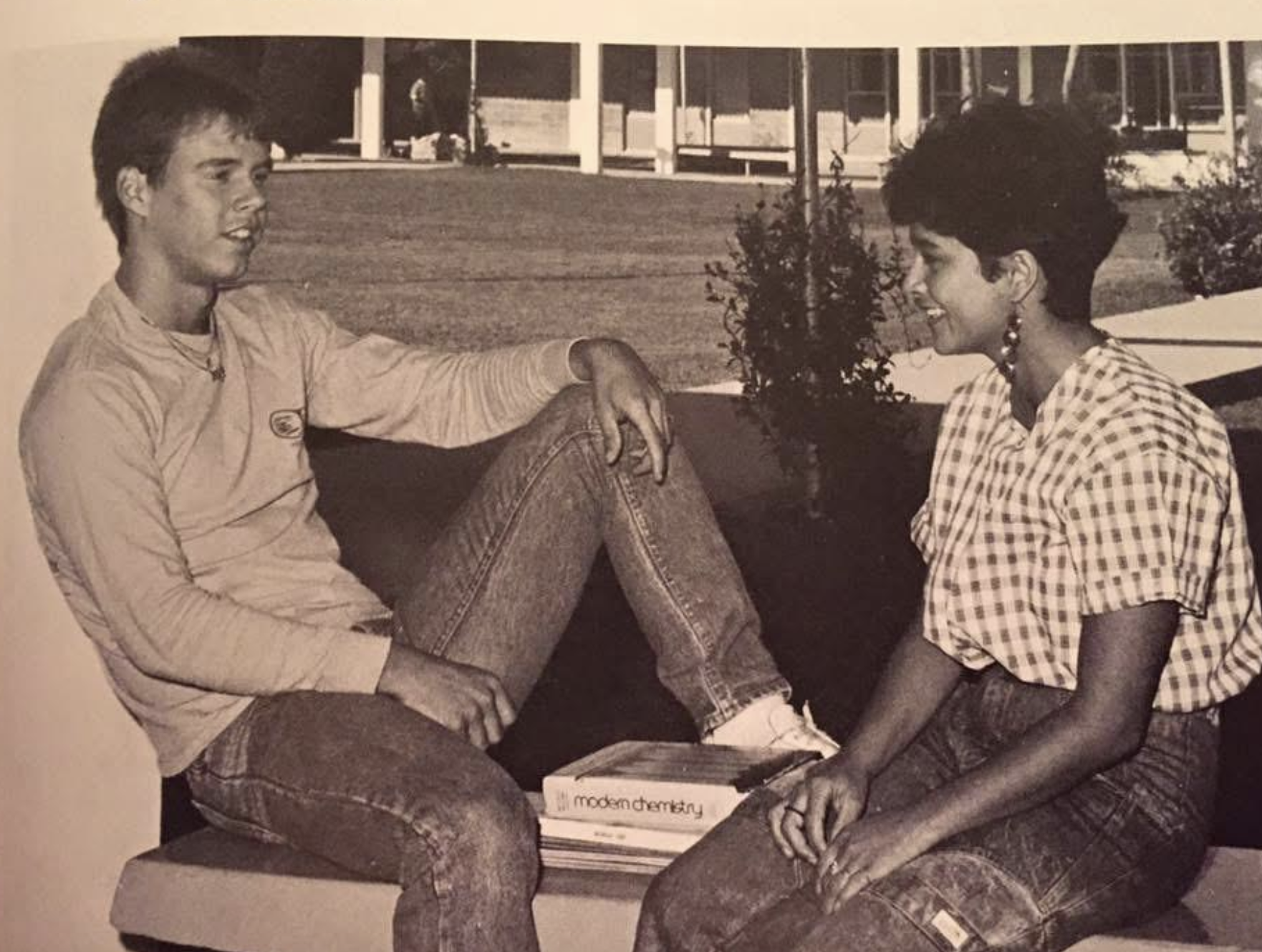 A young man and woman sit facing each other on a bench outdoors, with a textbook titled Modern Chemistry placed between them.