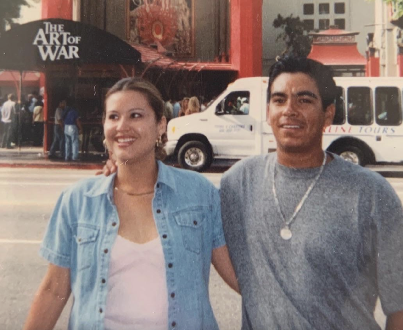 A smiling man and woman pose for a photo in front of a building with a sign that reads “The Art of War” and a parked white tour van.